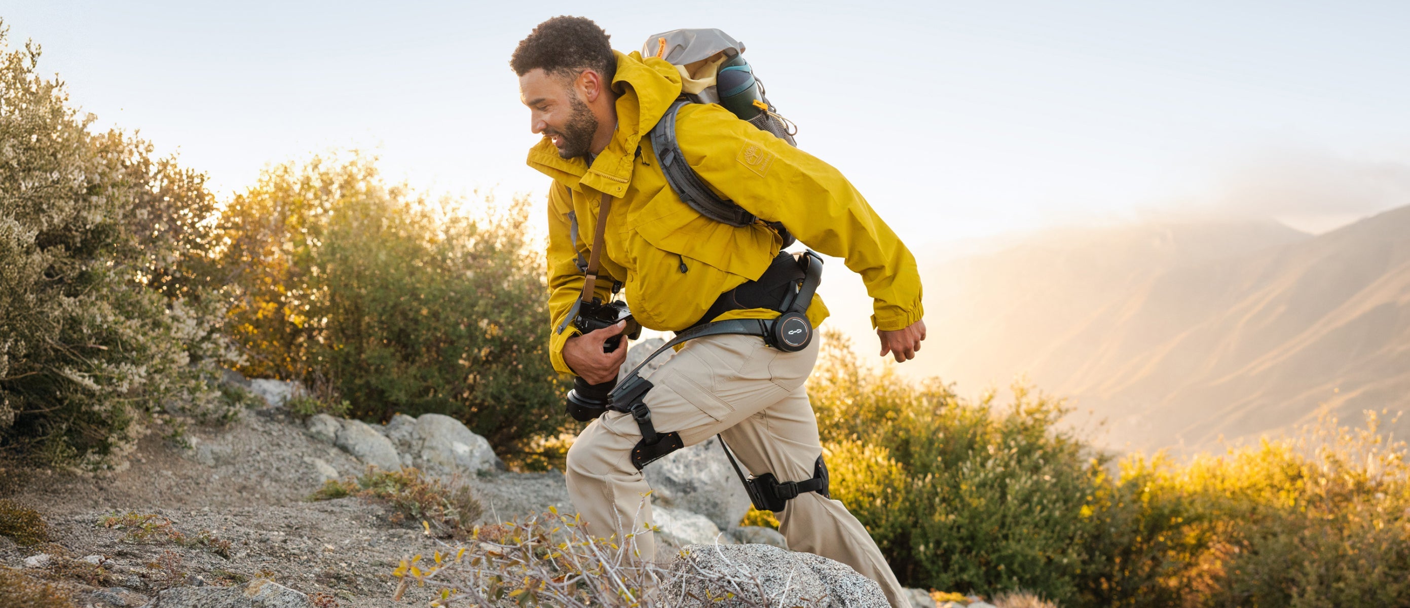 Outdoor photographer wearing a Hypershell X Series S exoskeleton and carrying equipment on a rocky trail